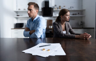 A man and woman sit at a table with crossed arms, facing away from each other. Documents related to their uncontested divorce and two wedding rings are placed on the table in the foreground.