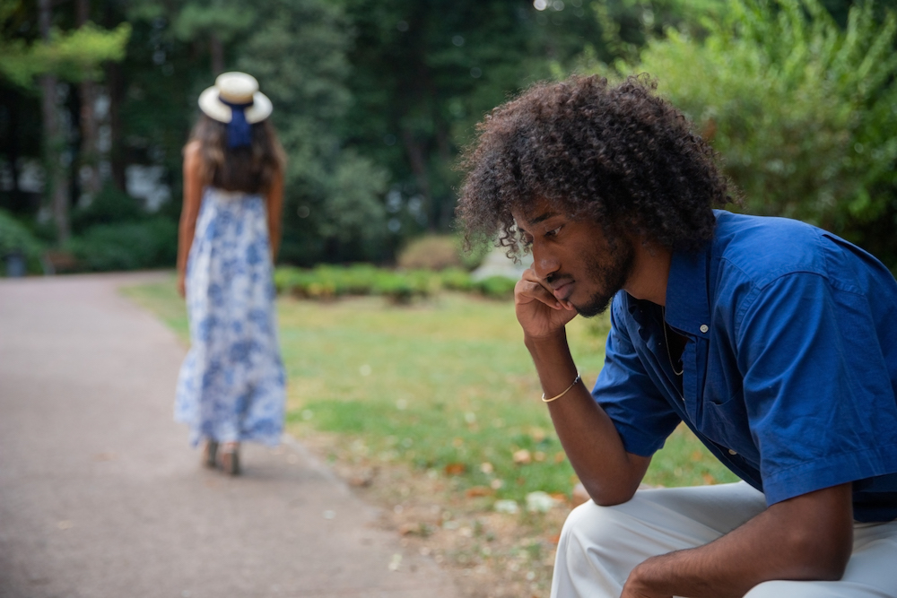 A man sits on a bench looking thoughtful while a woman in a blue dress walks away on a park path, reflecting the emotions often faced during divorce in Georgia.