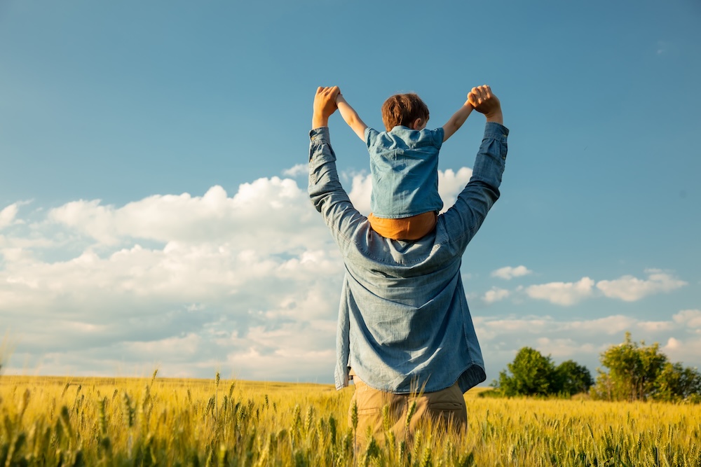 An adult carries a child on their shoulders in a field under a blue sky, both facing away from the camera with arms raised.